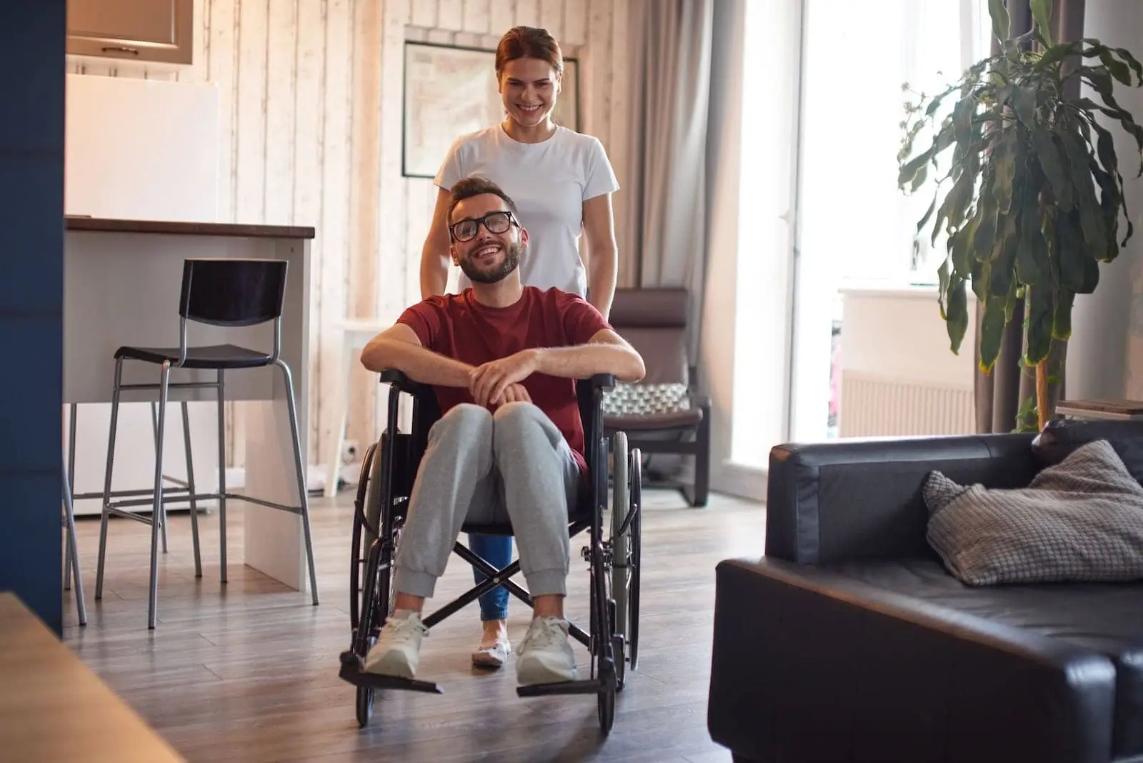 Junger Mann im Rollstuhl in seiner Wohnung, eine Begleiterin hinter ihm