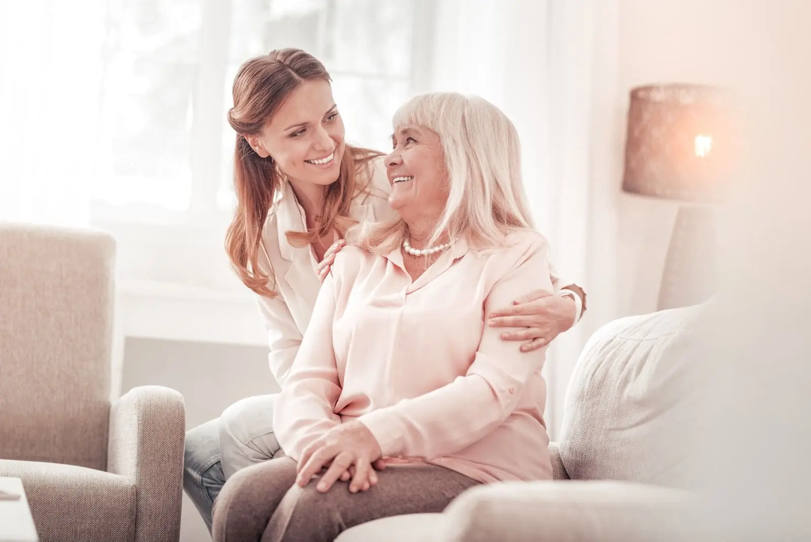 Jüngere Frau legt lachend den Arm um eine ältere Dame auf dem Sofa
